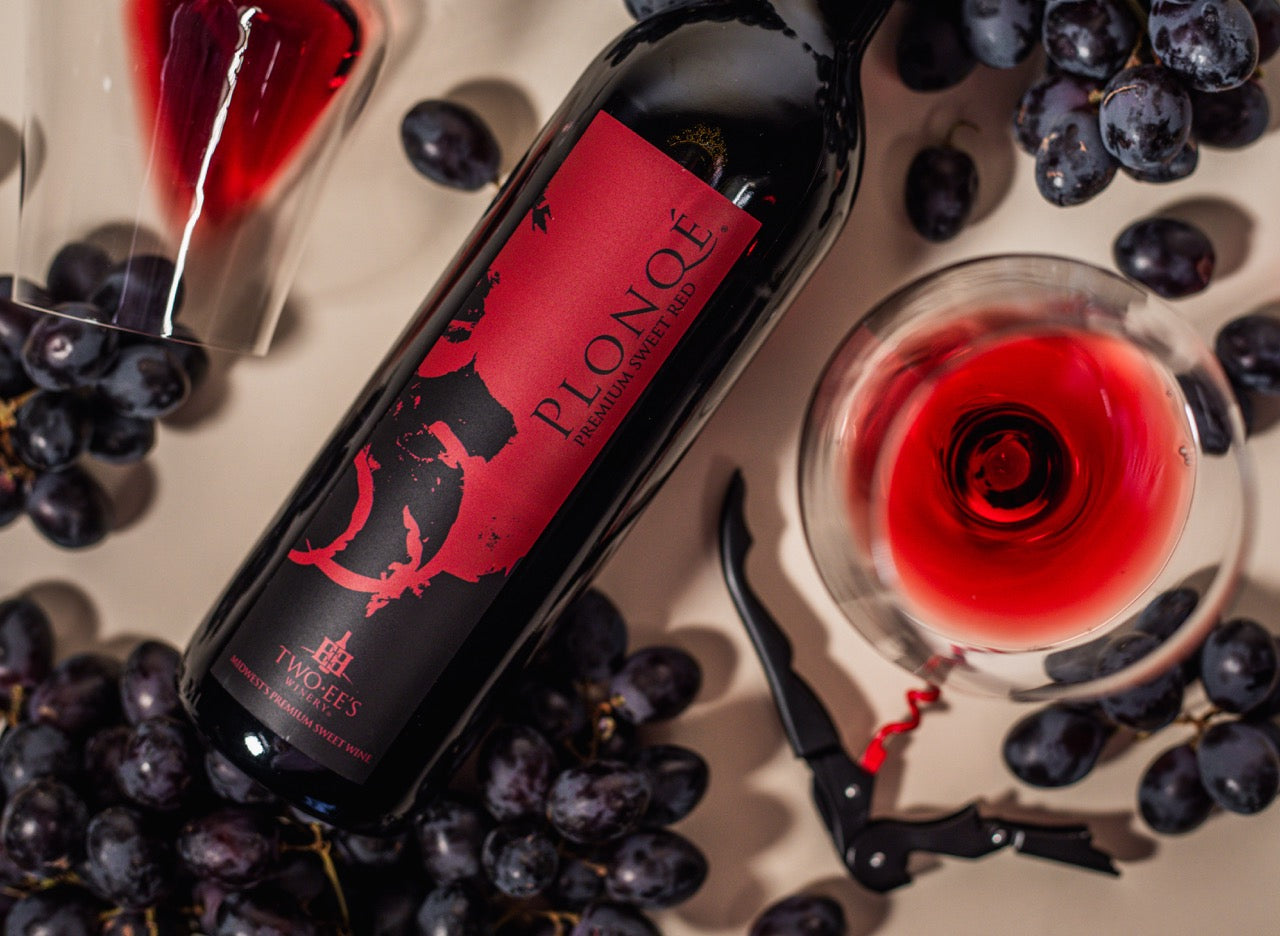 Bottle of Plonqé wine with a glass of red wine surrounded by grapes on a wooden surface.